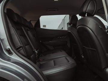 De afbeelding toont het interieur van een middelgrote SsangYong Korando e-Motion met zwartleren stoelen, gezien vanuit een open passagiersdeur. De stoelen zijn voorzien van veiligheidsgordels en hoofdsteunen en de auto oogt schoon en modern.