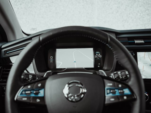 Close-up van het stuur van de SsangYong Korando e-Motion met een centraal digitaal display met navigatie-aanwijzingen en een ander scherm aan de rechterkant met een kaart. Zwart interieur met diverse knoppen op het stuur.