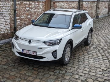 Een witte SsangYong Korando e-Motion met schuifdak en blauwe zijspiegelkappen staat geparkeerd op een geplaveide straat voor een bakstenen muur. Het voertuig heeft Europese kentekenplaten.