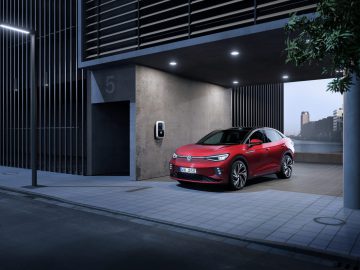 Een rood Volkswagen ID.5 elektrisch voertuig staat in de schemering geparkeerd onder een moderne carport met een laadstation in een stadsstraat.