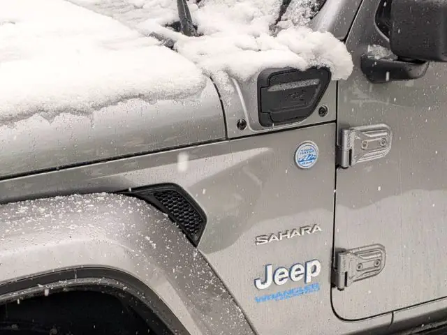 Close-up van een zilveren Jeep Wrangler Sahara, gedeeltelijk gehuld in sneeuw, geparkeerd op een besneeuwde straat.