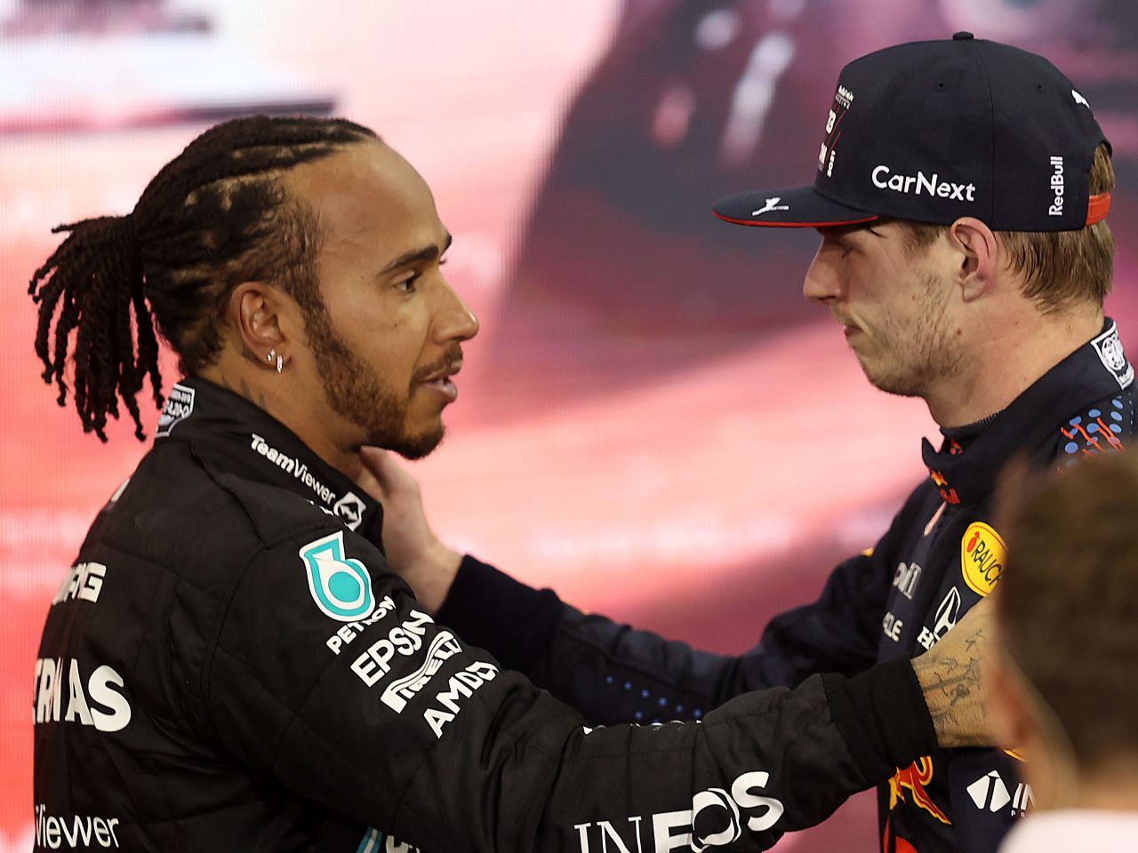 Twee autocoureurs in uniform en pet staan tegenover elkaar en wisselen een handdruk uit voor een strak Mercedes F1-voertuig.