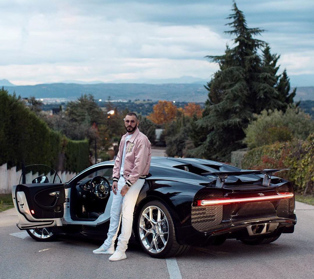 Een persoon staat naast een slanke, zwarte sportwagen met de deur aan de bestuurderszijde open op een schilderachtige weg met bomen en heuvels op de achtergrond, die doet denken aan de luxe voertuigen waar Karim Benzema de voorkeur aan gaf.