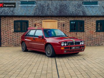 Een rode Lancia Delta Integrale HF Evolution staat geparkeerd voor een bakstenen gebouw met een houten deur en ramen met groene kozijnen. De auto is voorzien van veelkleurige racestrepen en een DK Engineering-kentekenplaat, die het race-erfgoed van de iconische Lancia Delta Integrale laat zien.