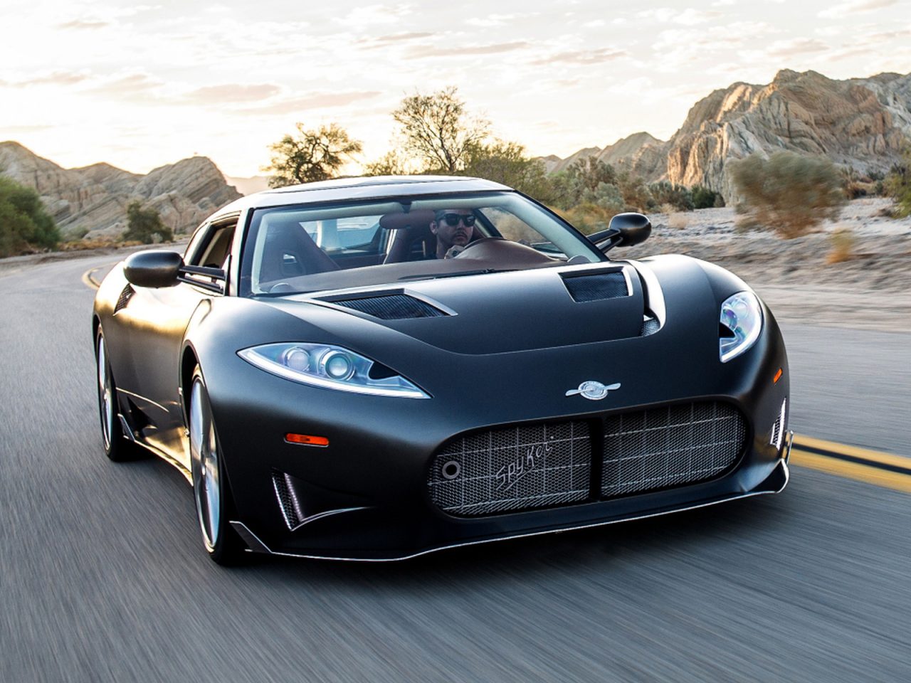 Een slanke, zwarte Spyker-sportwagen met een laag profiel rijdt op een woestijnweg met rotsachtige heuvels op de achtergrond.