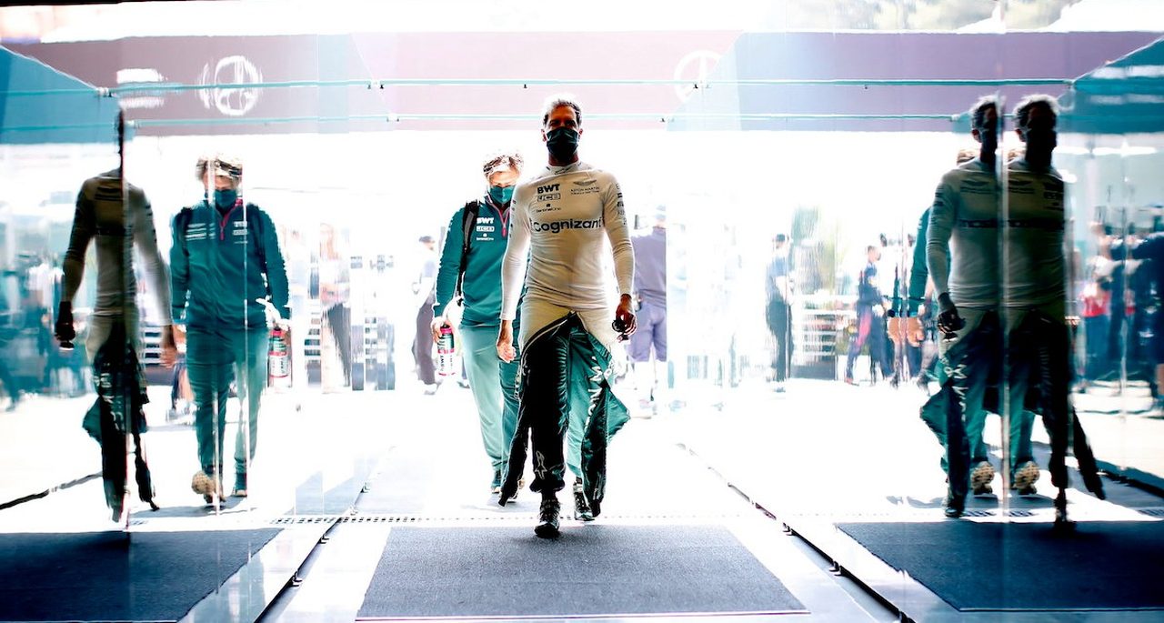 Twee coureurs in witte racepakken, één scherp in beeld, lopen door een glazen tunnel met reflecties aan beide kanten terwijl anderen op de achtergrond een race-evenement volgen, wat doet denken aan Verstappens vastberaden stappen voor een race.