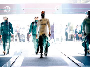 Twee coureurs in witte racepakken, één scherp in beeld, lopen door een glazen tunnel met reflecties aan beide kanten terwijl anderen op de achtergrond een race-evenement volgen, wat doet denken aan Verstappens vastberaden stappen voor een race.