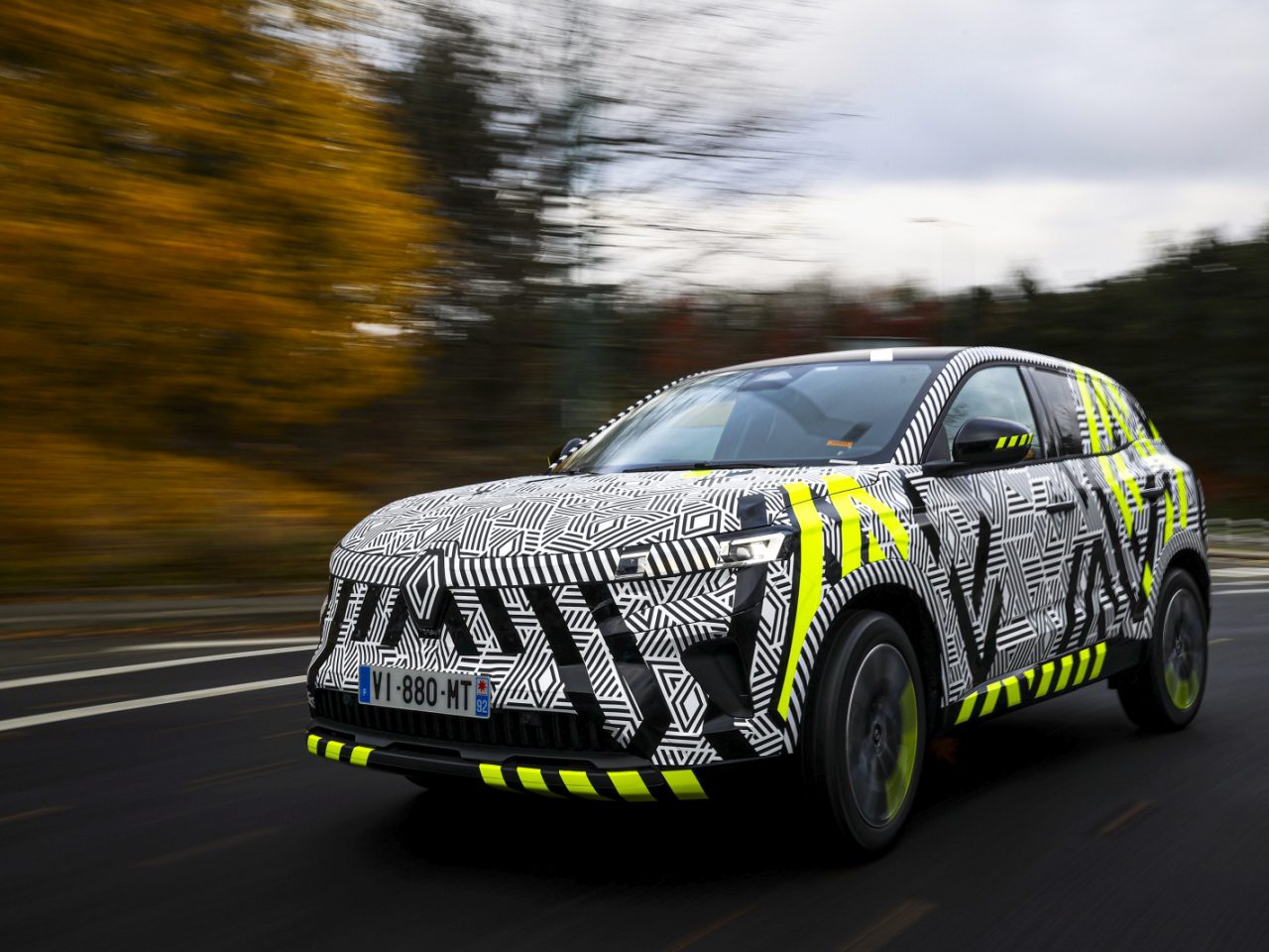 Een Renault Austral met een zwart-wit-geel camouflagepatroon rijdt over een weg met wazig herfstgebladerte op de achtergrond.