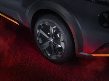 Close-up van een zwart Nissan Juke-wiel met strakke lichtmetalen velgen op een rood verlicht oppervlak. Het lijkt erop dat de auto geparkeerd staat, waarbij rood licht delen van de band en de grond benadrukt.