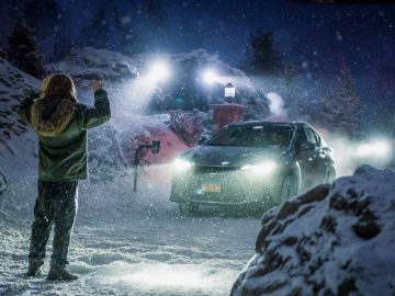 Een persoon in een winterjas en capuchon staat 's nachts op een besneeuwde weg, tegenover een naderende Lexus met koplampen aan. Met sneeuw bedekte rotsen en bomen omringen het tafereel.