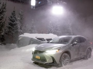 Een zilveren Lexus-SUV staat 's nachts geparkeerd op een besneeuwde weg, met een met sneeuw bedekte, omgevallen auto vlakbij. Felle lichten verlichten het tafereel en op de achtergrond zijn hoge, met sneeuw bedekte bomen zichtbaar.