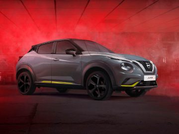 Een grijze Nissan Juke met zwarte wielen en gele accenten staat geparkeerd in een industriële omgeving, gehuld in rode verlichting en mistige mist.