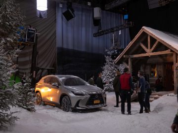 Mensen zijn bezig met het opzetten van een besneeuwde scène op een filmset. Een zilverkleurige Lexus-SUV staat geparkeerd tussen kunstsneeuw en bomen in de buurt van een houten constructie, terwijl bemanningsleden eromheen werken.