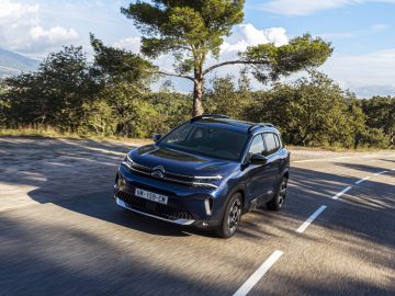 Een blauwe Citroën C5 Aircross 2022 rijdt op een schilderachtige, bochtige weg omringd door bomen en heuvels onder een heldere hemel.