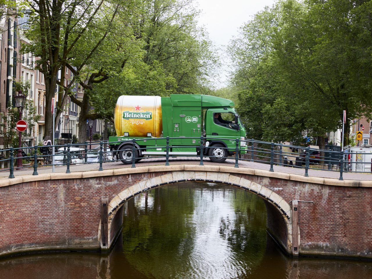 Een groene Heineken-bestelwagen, beladen met autonoom tankbier voor de Amsterdamse horeca, staat geparkeerd op een stenen brug over een gracht, omgeven door bomen en gebouwen. Het tafereel weerspiegelt perfect de essentie van 100% groen bezorgen in hartje Amsterdam.