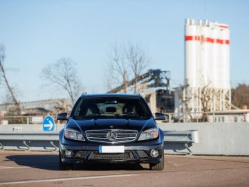 Een zwarte Mercedes-Benz-auto, die doet denken aan die van racelegende Michael Schumacher, staat geparkeerd op een industriegebied met een gedeeltelijk wazige fabriek op de achtergrond onder een heldere hemel.