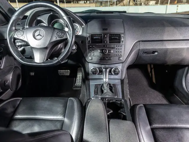 Binnenaanzicht van een auto met de bestuurdersstoel, het stuur met Mercedes-Benz-logo, het dashboard met bedieningselementen en de middenconsole met versnellingspook. Zwart lederen stoelen en moderne designkenmerken geïnspireerd op de precisie en elegantie van de race-erfenis van Michael Schumacher.