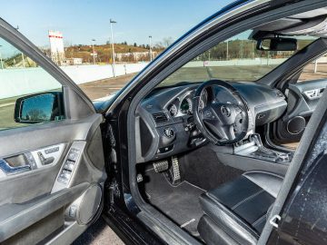 Interieur van een zwarte auto met de deur aan de bestuurderszijde open, met het stuur, het dashboard en de middenconsole met versnellingspook; op de achtergrond zijn een parkeerplaats en een gebouw zichtbaar. Je zou je bijna kunnen voorstellen dat Michael Schumacher zich voorbereidt om het stuur over te nemen.