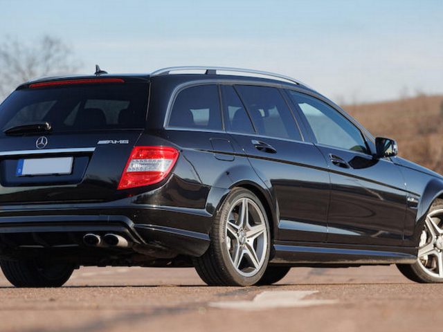 Een zwarte Mercedes-Benz AMG-stationwagen, die doet denken aan iets waarin Michael Schumacher zou kunnen rijden, staat op een heldere dag geparkeerd op een open plek, gezien vanaf de achterkant en de linkerkant.