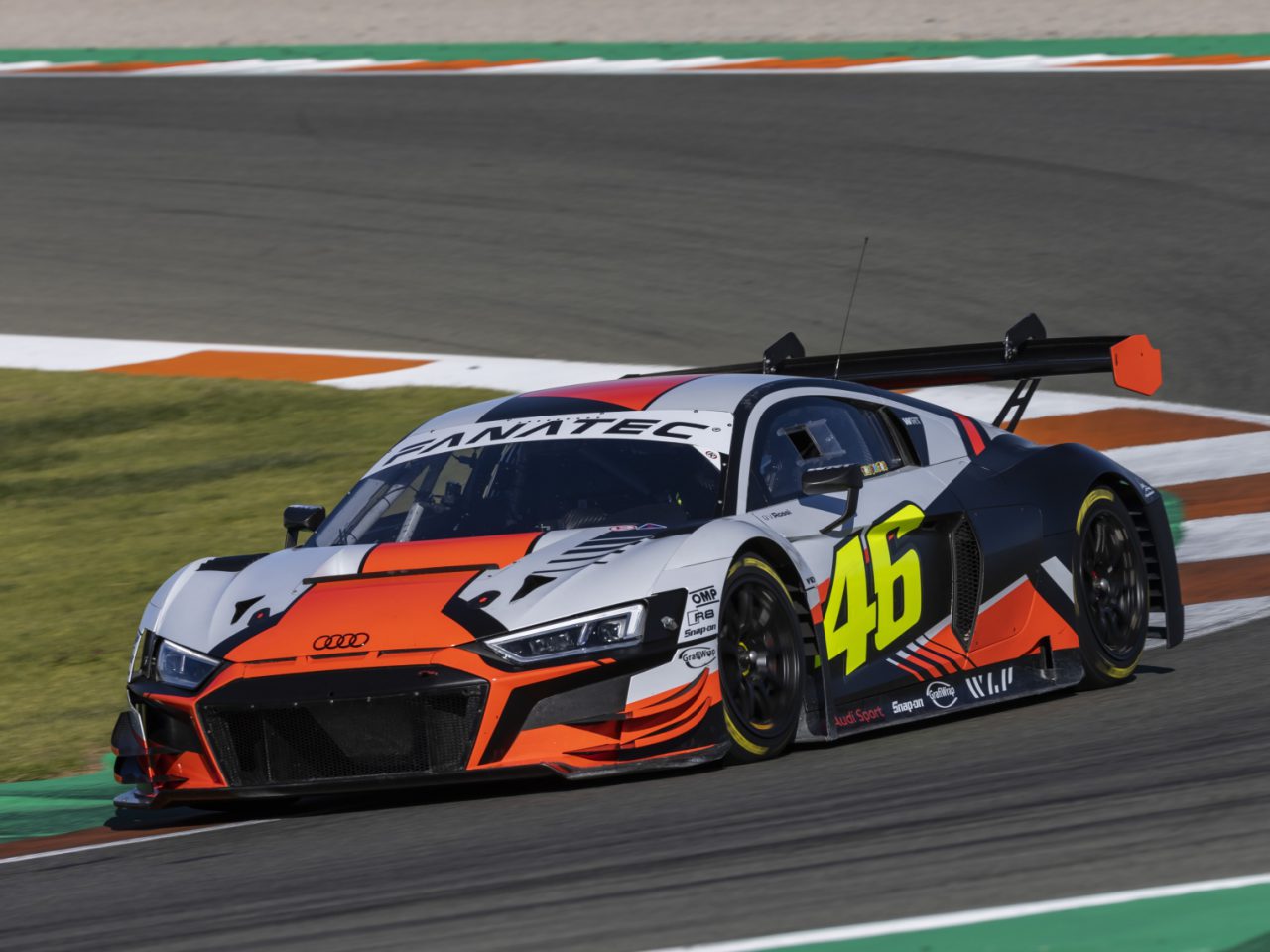 Een racewagen met het nummer 46 op de zijkant, een weerspiegeling van het legendarische nummer van Valentino Rossi, heeft een zwart, wit en oranje ontwerp terwijl hij tijdens een race overdag over het circuit snelt.