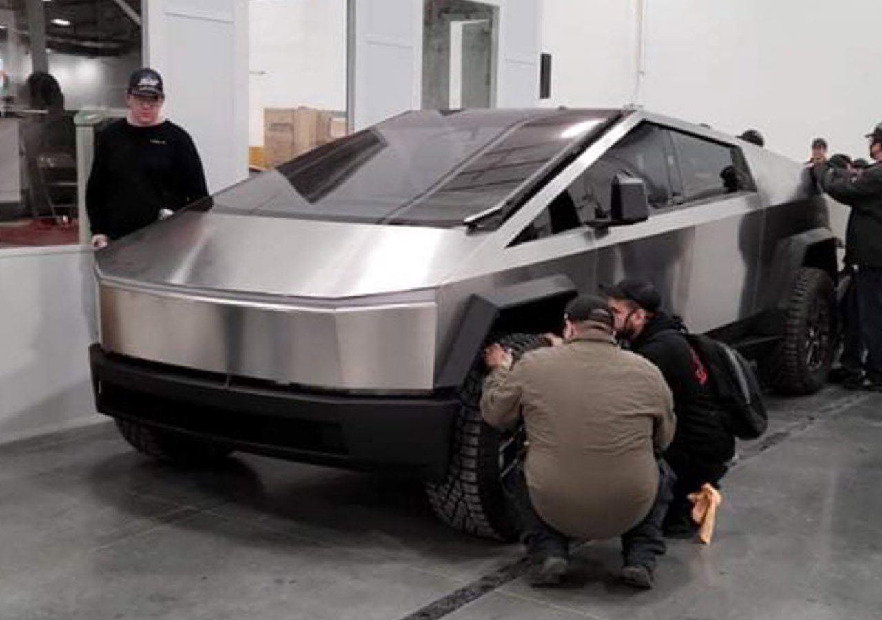 Werknemers verzamelen zich rond een futuristische roestvrijstalen vrachtwagen die lijkt op een Tesla Cybertruck in een industriële omgeving. Sommigen inspecteren en passen het voertuig aan.