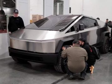 Werknemers verzamelen zich rond een futuristische roestvrijstalen vrachtwagen die lijkt op een Tesla Cybertruck in een industriële omgeving. Sommigen inspecteren en passen het voertuig aan.