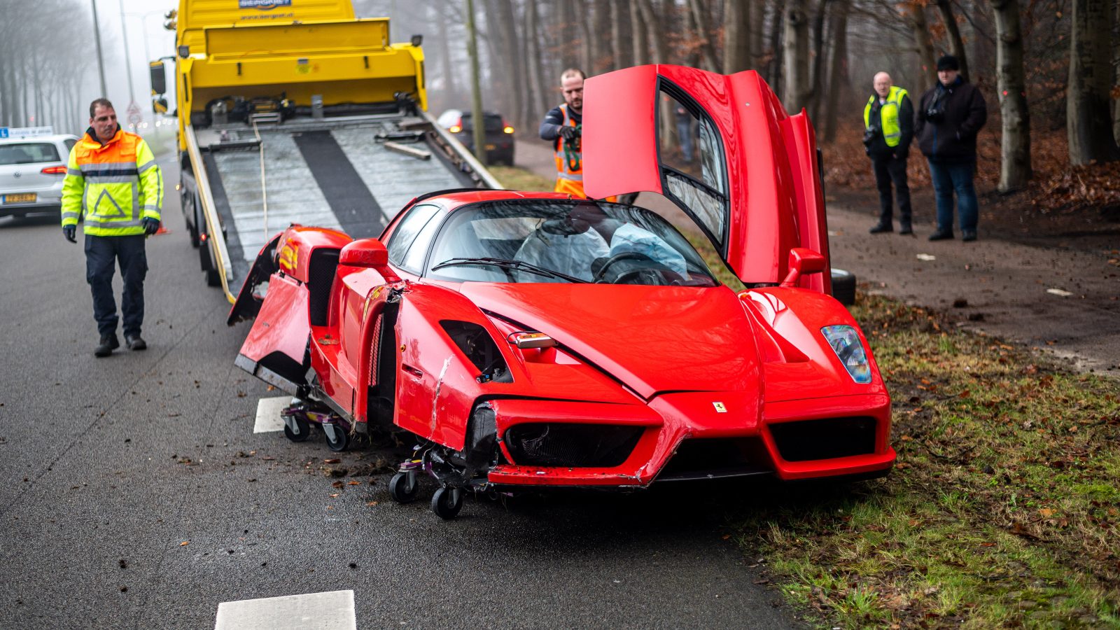 Een beschadigde rode Ferrari wordt na een crash op een takelwagen aan de kant van de weg geladen. Hulpdiensten staan in de buurt en de auto heeft aanzienlijke schade aan de voorkant.