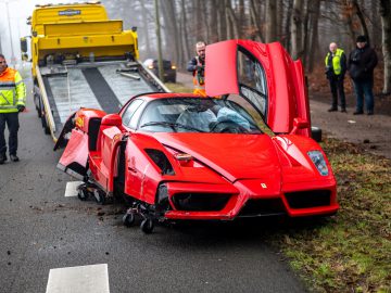 Een beschadigde rode Ferrari wordt na een crash op een takelwagen aan de kant van de weg geladen. Hulpdiensten staan in de buurt en de auto heeft aanzienlijke schade aan de voorkant.