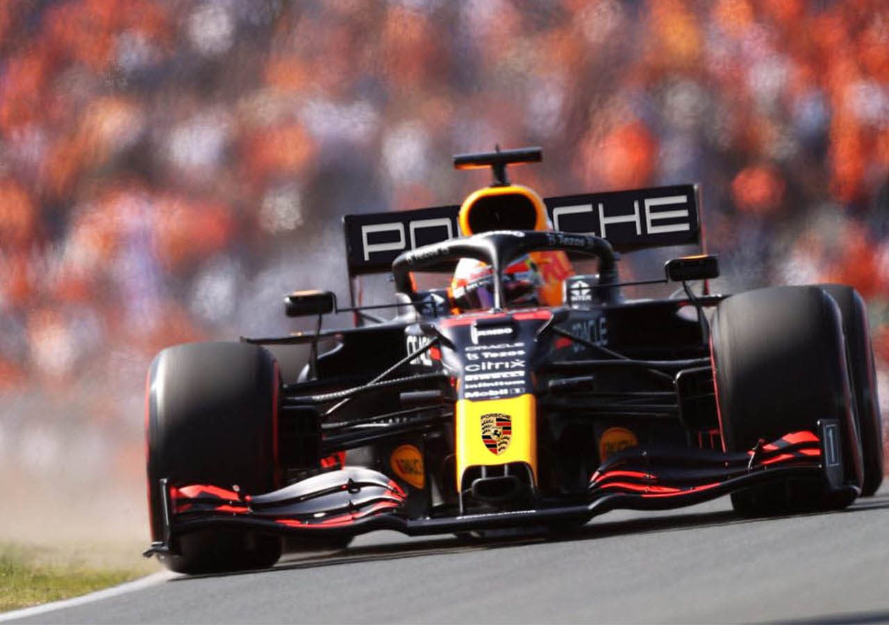 Een Formule 1-auto met het Porsche-logo op de achtervleugel racet op een circuit met een wazige menigte op de achtergrond, terwijl een Audi Formule 1-auto hem op de voet volgt.