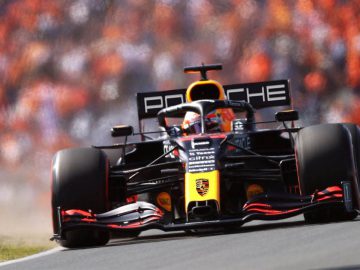 Een Formule 1-auto met het Porsche-logo op de achtervleugel racet op een circuit met een wazige menigte op de achtergrond, terwijl een Audi Formule 1-auto hem op de voet volgt.