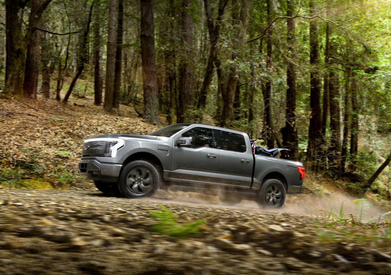 Een grijze Ford F-150 rijdt over een onverharde weg door een bos met aan weerszijden bomen en bladeren.