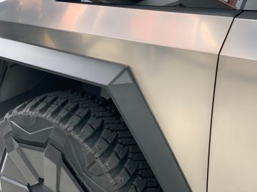 Close-up van het achterwiel en het hoekige metalen frame van een futuristisch voertuig, met het robuuste bandenontwerp dat doet denken aan de Tesla Cybertruck.