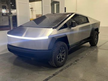 Een futuristische, vierkante Tesla Cybertruck met een hoekig ontwerp en metallic afwerking staat geparkeerd in een industrieel gebouw.