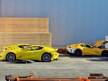 Twee gele Ferrari-sportwagens met zichtbare schade staan geparkeerd op een industrieterrein nabij gestapelde houten pallets. De ene auto heeft de motorkap open, terwijl de deur van de andere auto op een kier staat.