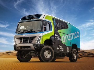 Een robuust elektrisch terreinvoertuig met het opschrift ‘Aramco’ staat in een woestijnlandschap met helderblauwe luchten en belichaamt de geest van de Dakar Rally. Het voertuig is opgetild en voorzien van grote, stevige banden geschikt voor ruw terrein.