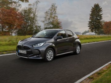Een donkergrijze Mazda 2 Hybrid rijdt over een landelijke weg omzoomd door grasvelden en bomen met herfstbladeren onder een bewolkte hemel.