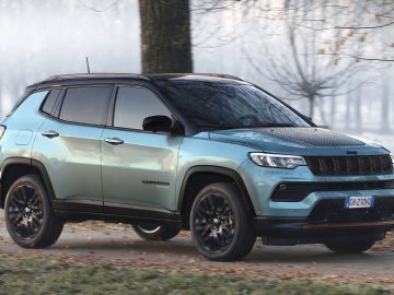 Een lichtblauwe Jeep SUV rijdt op een mistige weg omzoomd met bomen, voorzien van zwarte accenten en een modern design.