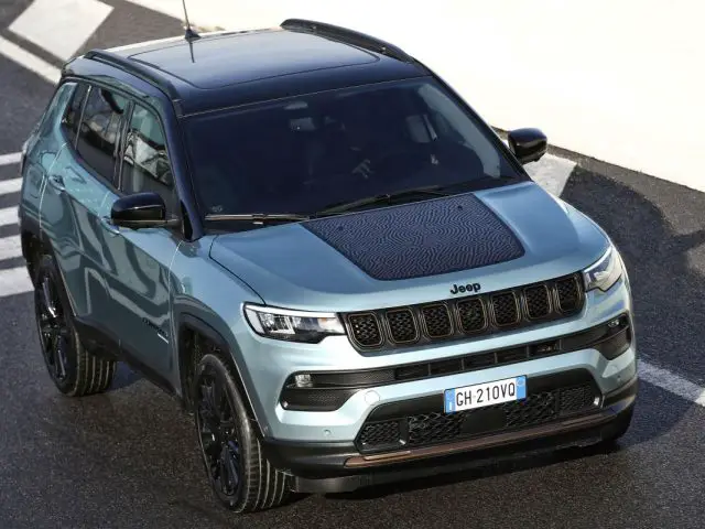 Een blauwgroenkleurige Jeep SUV met een zwart dak, geparkeerd op een verhard oppervlak met witte markeringen en een Italiaans kenteken "GH 210VO" zichtbaar.