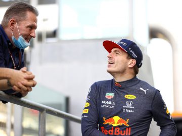 Twee mannen zijn aan het praten; de een draagt een Red Bull Racing-uniform en pet met de naam van Verstappen op de achterkant, terwijl de ander op een metalen reling leunt met een gezichtsmasker onder zijn kin.