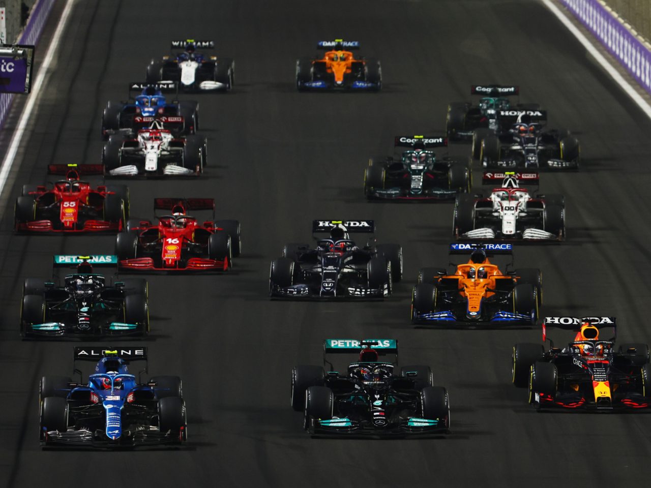 Een groep Formule 1-auto's met verschillende teamkleuren racet 's nachts op een circuit, elk met hun unieke Startnummer, opgesteld in meerdere rijen.