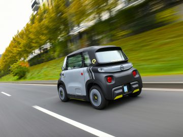 Een klein, grijs en zwart tweezits elektrische Opel Rocks-e-voertuig rijdt over een stadsweg met groene struiken en gebouwen op de achtergrond.