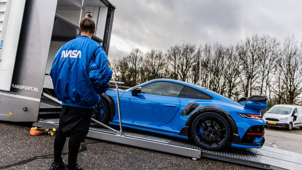 Een persoon in een NASA-jasje kijkt toe hoe een blauwe La Fuente-auto op een bewolkte dag op een transportwagen wordt geladen.