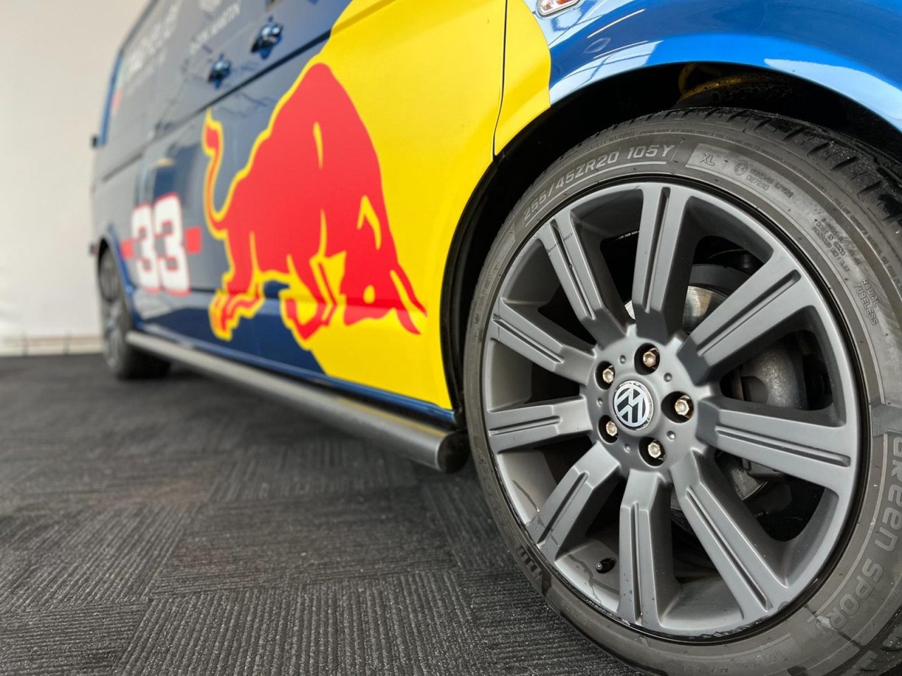 Close-up van het linker voorwiel van een auto met grijze velg, voorzien van een kleurrijke voertuigwrap met een Red Bull-afbeelding en het nummer 33. De band, met het merk "PIRELLI P ZERO NERO GT", verwijst naar het racevermogen van Max Verstappen.