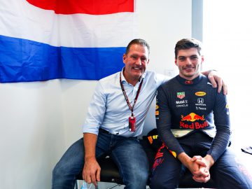 Twee mannen zitten naast elkaar in een kamer, waarbij de één een Red Bull-racepak draagt dat fans vaak associëren met Verstappen, en de ander in vrijetijdskleding. Aan de muur achter hen hangt een vlag met rode, witte en blauwe strepen.