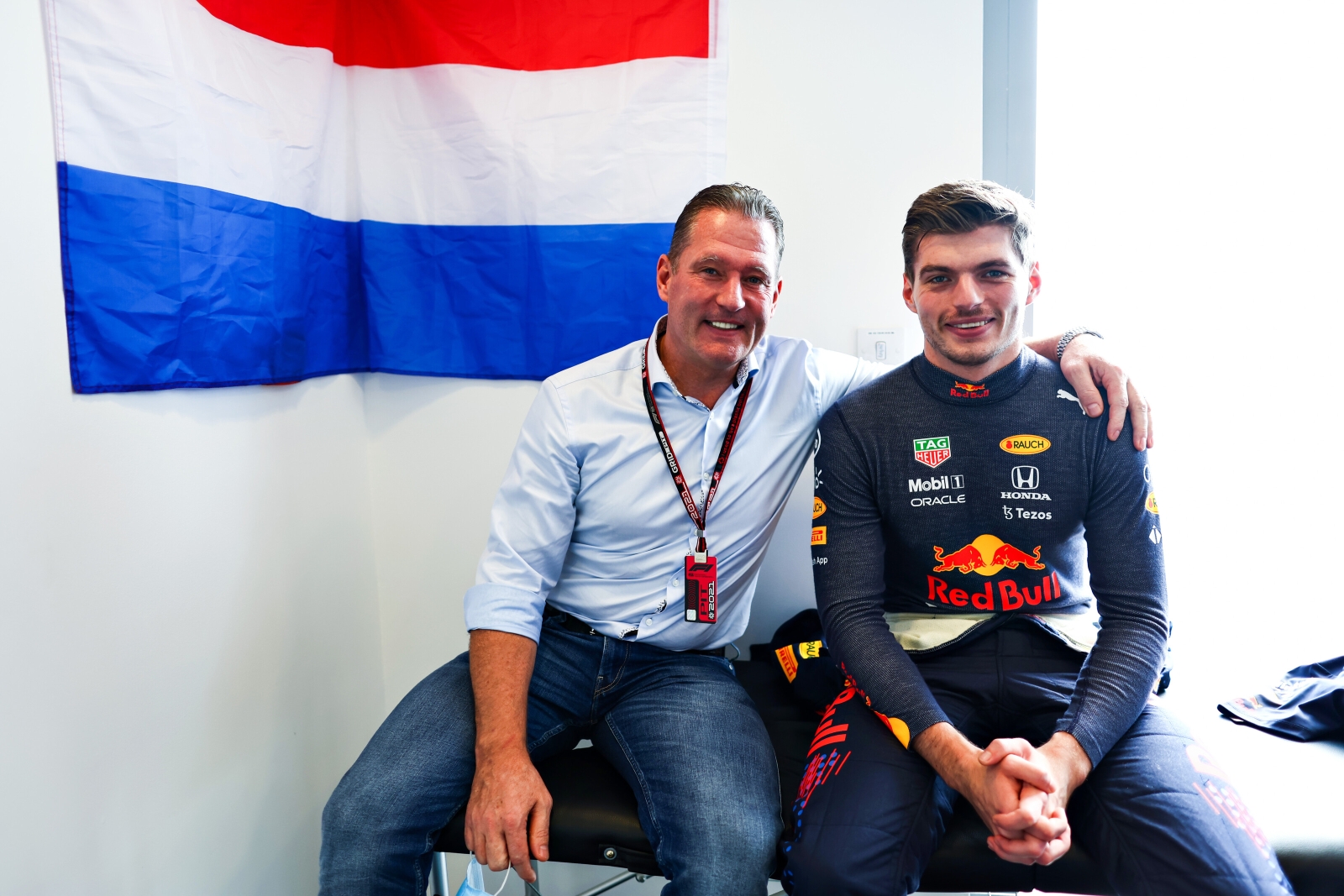 Twee mannen zitten naast elkaar in een kamer, waarbij de één een Red Bull-racepak draagt dat fans vaak associëren met Verstappen, en de ander in vrijetijdskleding. Aan de muur achter hen hangt een vlag met rode, witte en blauwe strepen.
