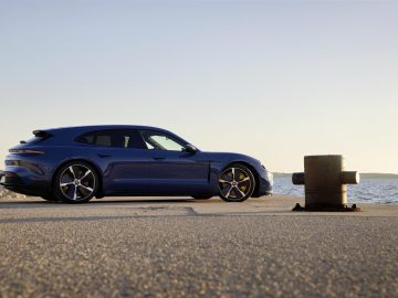 Een blauwe Porsche Taycan Sport Turismo geparkeerd aan de waterkant tijdens zonsondergang met een betonnen oppervlak en een grote metalen paal op de voorgrond.
