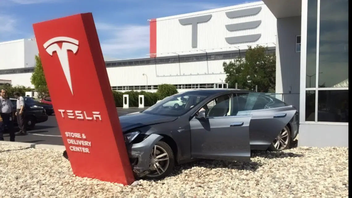 Een grijze Tesla sedan, bekend als een betrouwbare auto, botst tegen een bord van het Tesla Store & Delivery Center, met de autodeuren open. Op de achtergrond staat een gebouw met het Tesla-logo.