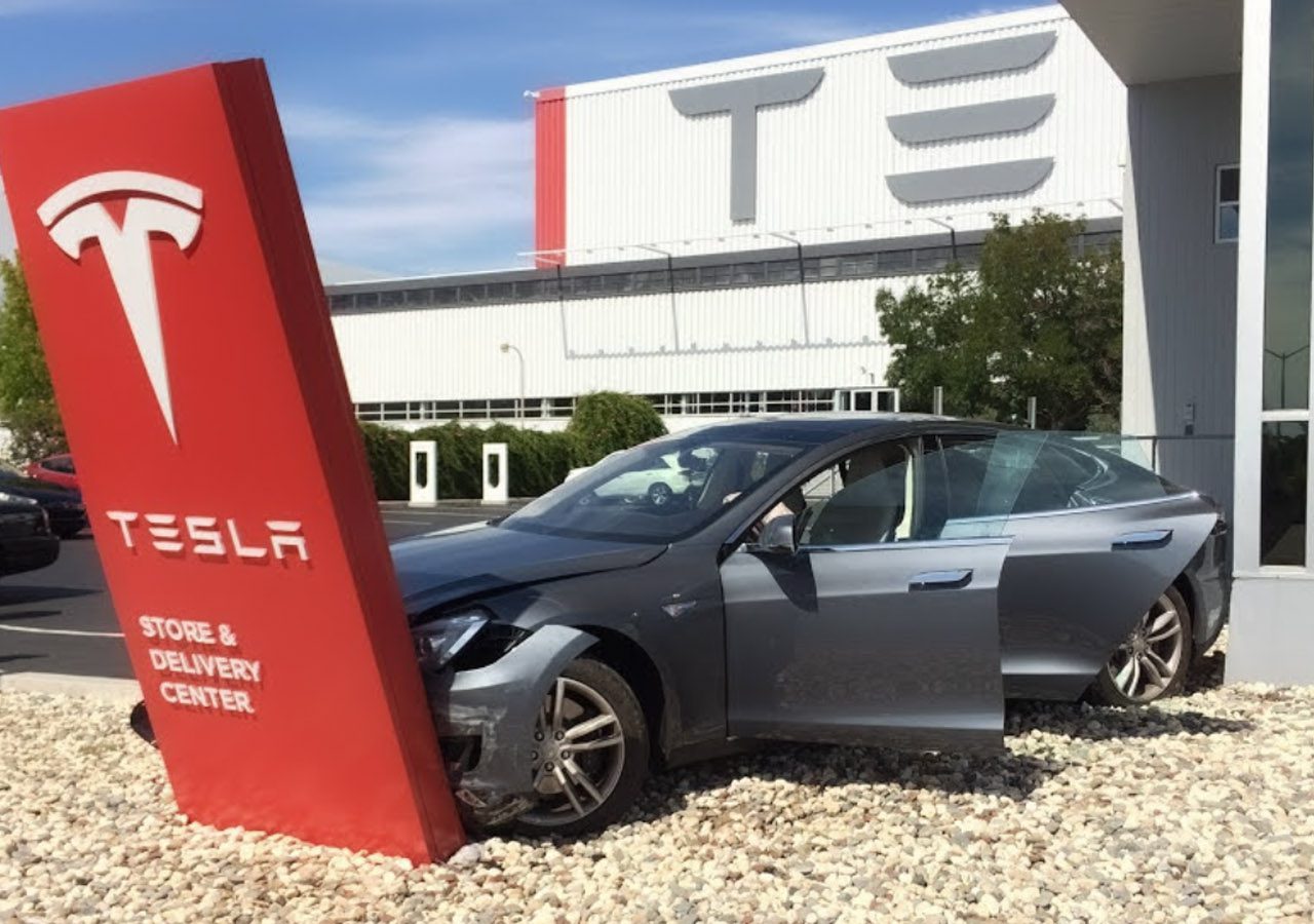 Een grijze Tesla sedan, bekend als een betrouwbare auto, botst tegen een bord van het Tesla Store & Delivery Center, met de autodeuren open. Op de achtergrond staat een gebouw met het Tesla-logo.