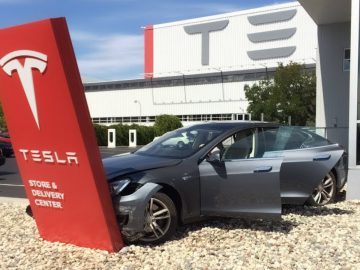 Een grijze Tesla sedan, bekend als een betrouwbare auto, botst tegen een bord van het Tesla Store & Delivery Center, met de autodeuren open. Op de achtergrond staat een gebouw met het Tesla-logo.