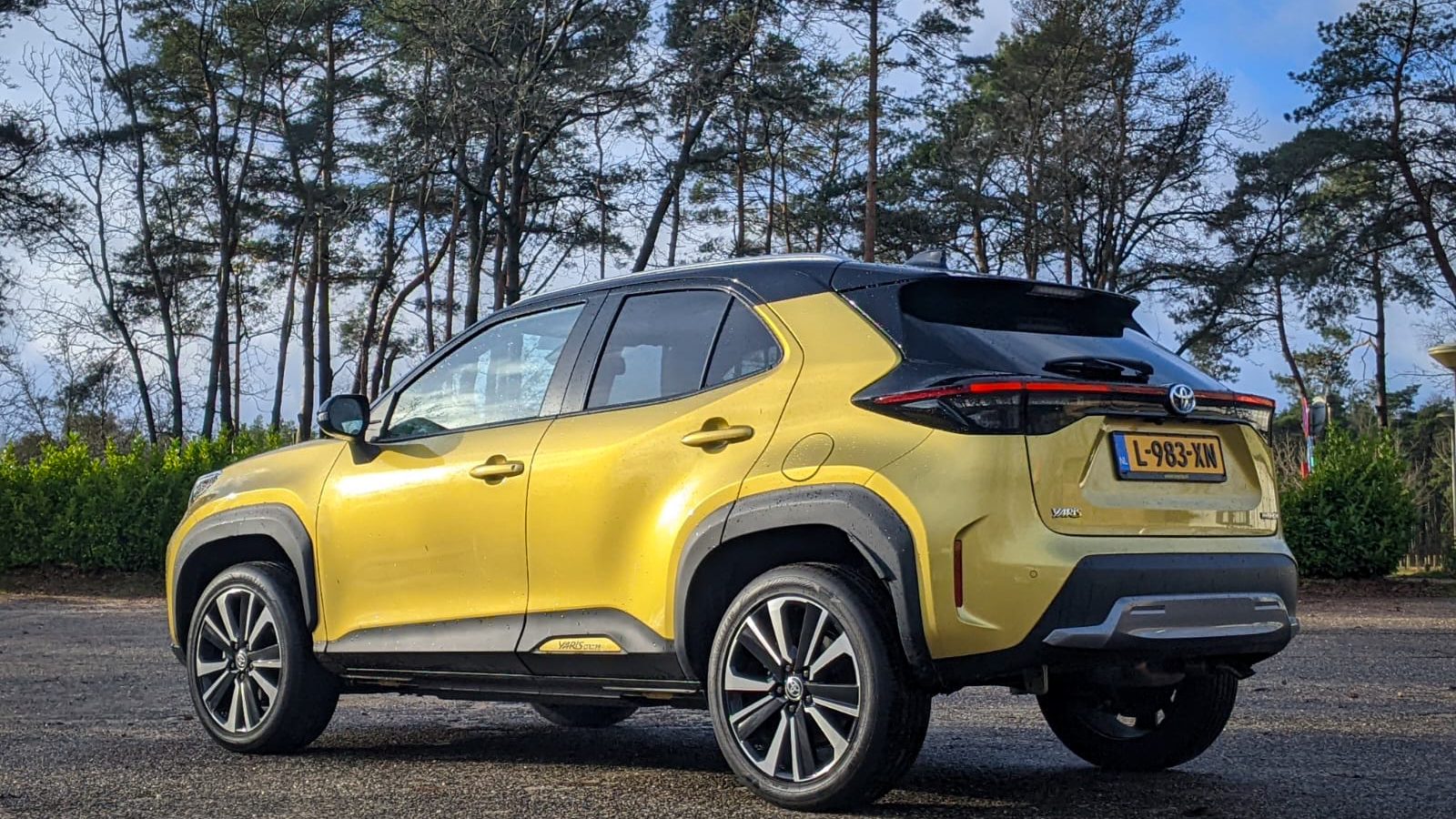 Een gele en zwarte Toyota Yaris Cross compacte SUV staat geparkeerd op een buitenterrein met op de achtergrond bomen en een bewolkte lucht. De auto heeft een Nederlands kenteken.